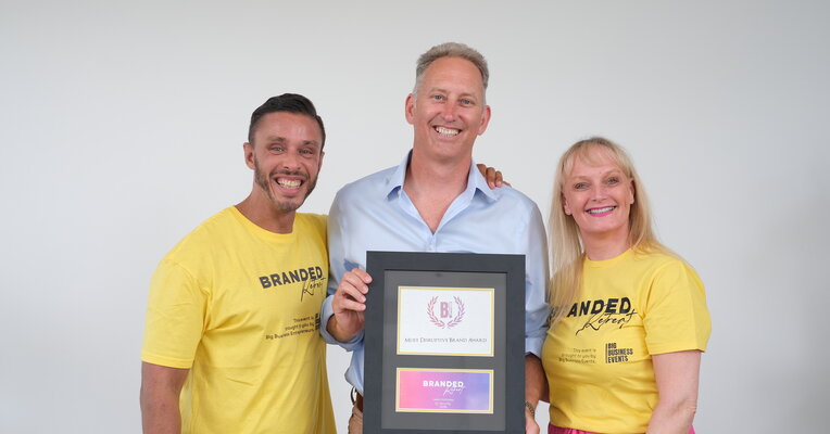 Jason Holloway of QL Security holding the Most Disruptive Brand Award, flanked by the two Branded Retreat hosts